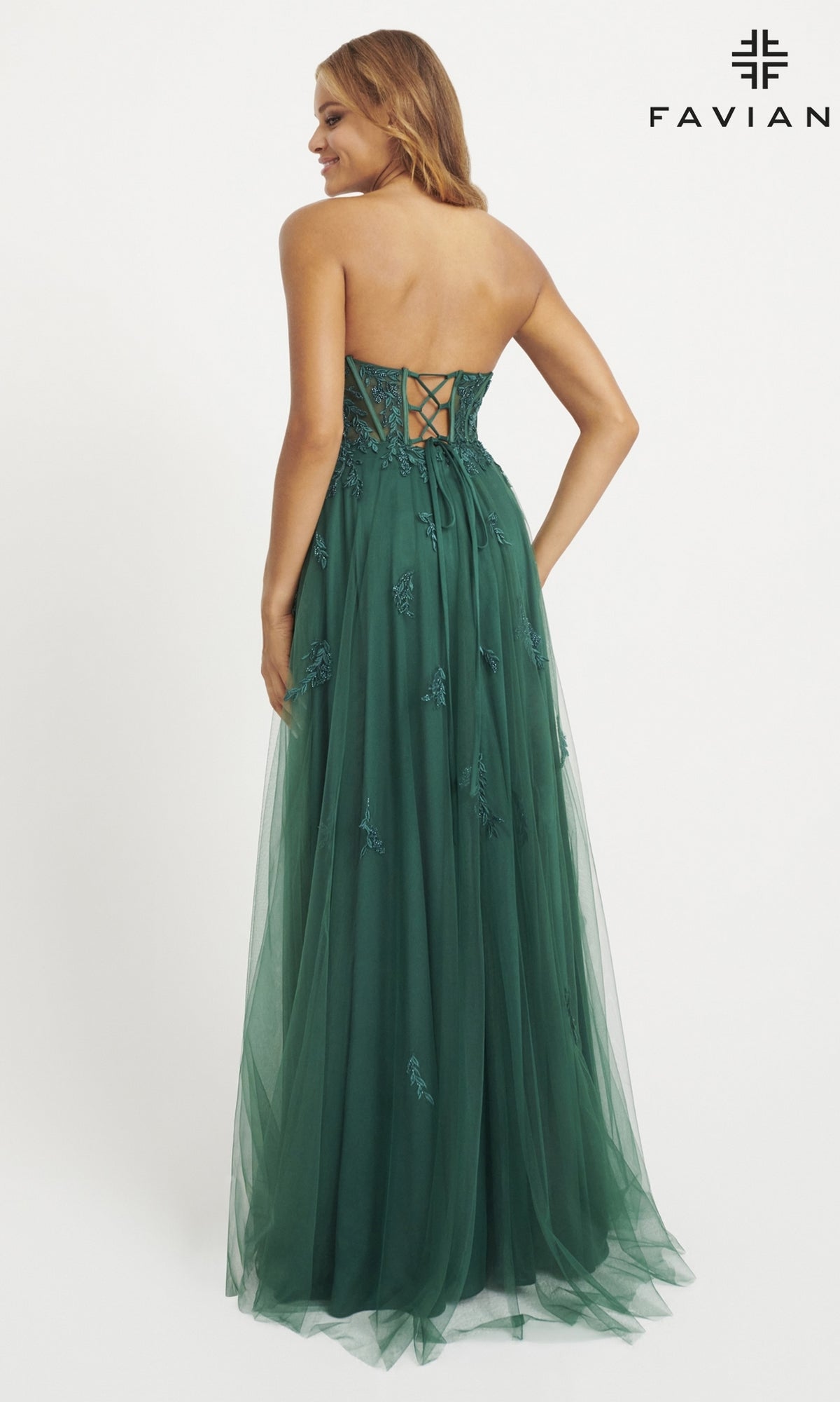 A woman stands against a plain white background wearing the Strapless Tulle Faviana Long Prom Dress 11057, a floor-length green gown with a lace-up corset bodice and floral embellishments.